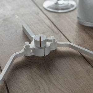 Puede incluir: Un objeto blanco de tres patas con un cubo cilíndrico central y una línea vertical marrón. Las patas se extienden hacia afuera, creando una forma de estrella. El objeto está sobre una superficie de madera, con un vaso y una taza en el fondo.