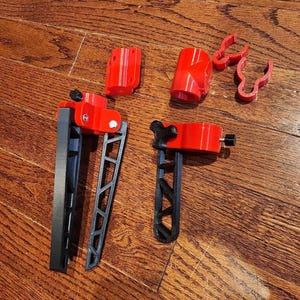 May include: Assortment of red and black 3D printed parts for a camera tripod. The parts include a base, a leg, a connector, and a clamp.