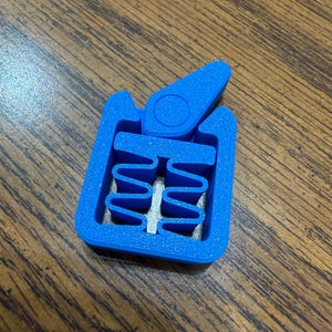 May include: A bright blue plastic clamp with a rectangular frame and a hinged lever. The interior features a wavy design. The clamp is designed to hold objects together. The item is on a wooden surface.