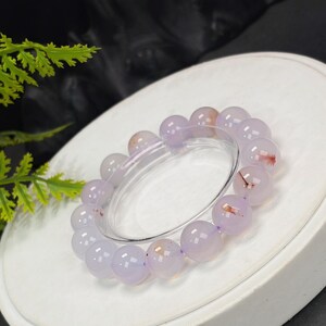 May include: A bracelet featuring round, polished beads in shades of lavender and pale peach. The beads are strung together, forming a circular shape. The bracelet is displayed on a white surface, with a clear elastic band visible.