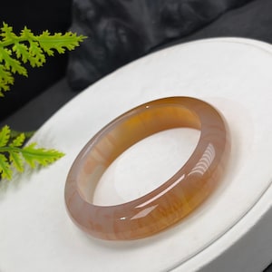 May include: A smooth, polished bangle bracelet crafted from translucent amber-toned agate stone. The circular jewelry piece features natural swirling patterns in shades of honey, gold, and light brown, with a glossy finish that reflects light.