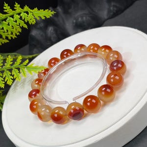Könnte beinhalten: Ein Armband mit polierten, runden Perlen in Orange- und Bernsteintönen. Die Perlen sind aufgefädelt und bilden ein kreisförmiges Design. Das Armband wird auf einer weißen Oberfläche präsentiert.