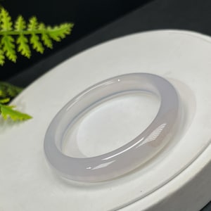 May include: A light gray, translucent jade bangle bracelet. The smooth, polished surface reflects light, showcasing the natural variations in the stone. The bracelet is a circular shape, designed to be worn on the wrist.