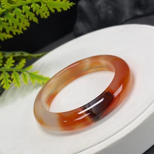 以下が含まれることがあります： 半透明のメノウ石で作られた、滑らかで磨き上げられたバングルブレスレット。このブレスレットは、クリームホワイトから鮮やかなオレンジ、そして深い赤褐色へと変化する温かみのある色のグラデーションが特徴で、はっきりとした暗い内包物が見られます。