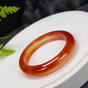 May include: A polished, translucent orange and white agate bangle bracelet. The bracelet has a smooth, rounded design and a glossy finish. The color transitions from a deep orange to a lighter, almost white hue. The bracelet is displayed on a white surface.