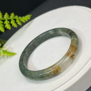 May include: A jade bangle bracelet with a mix of green, white, and brown hues. The bracelet is smooth and polished, with a circular shape. The colors blend together, creating a natural and organic look. The bracelet is a piece of jewelry.