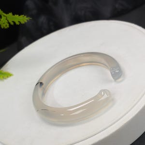 以下が含まれることがあります： 明るいグレーの、オープンエンドのカフブレスレット。ブレスレットは、微妙な色のバリエーションと滑らかで洗練された仕上げが施された半透明の素材で作られています。デザインはシンプルでエレガントです。