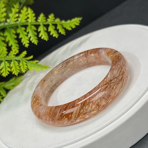 Può includere: Un braccialetto rigido traslucido, rosa chiaro e marrone con un aspetto unico, naturale e testurizzato. Il braccialetto è realizzato in un tipo di quarzo con inclusioni di fibre marroni dorate.