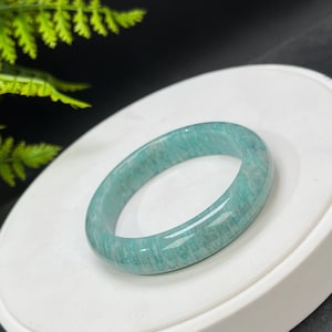 May include: A light green jade bangle bracelet with a smooth, polished finish. The bracelet is made of natural stone and has a delicate, feminine look.