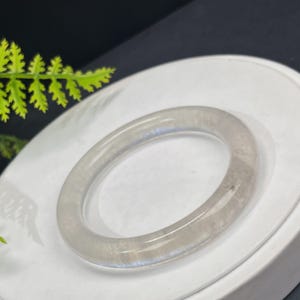 Pode incluir: Uma pulseira clara e translúcida com um design arredondado. A pulseira é de cor cinza claro e parece ser feita de um material liso e polido. A pulseira é circular e assenta numa superfície branca.