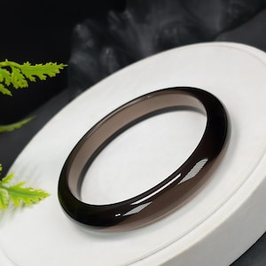 Könnte beinhalten: Ein poliertes, dunkelbraunes Edelstein-Armreif-Armband. Das glatte, oval geformte Armband wird auf einer weißen Oberfläche mit dunklem Hintergrund präsentiert. Das Armband hat eine glänzende Oberfläche, die das Licht reflektiert.