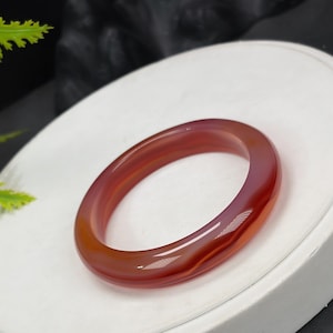 May include: A polished, translucent agate bangle bracelet. The bracelet features a gradient of colors, transitioning from a deep red to a lighter orange and pink hue. The smooth, rounded design of the bracelet is displayed on a white surface.