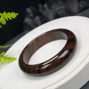 May include: A polished, dark brown wooden bangle. The bracelet has a smooth, glossy finish and a natural wood grain pattern. The circular bracelet is made from a solid piece of wood. The diameter is approximately 7 cm.