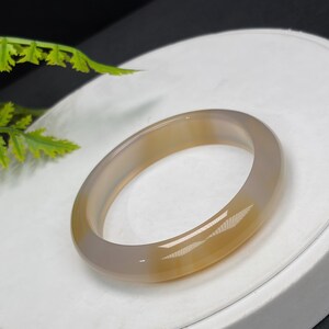 Può includere: Un braccialetto liscio e lucido nei toni del beige e del grigio. Il braccialetto circolare è realizzato in un materiale traslucido, con un effetto sfumato. È esposto su una superficie bianca, con una pianta verde sullo sfondo. Il diametro è di circa 6,5 cm.