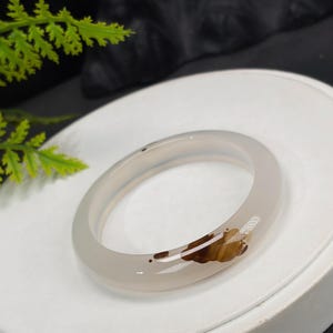 Può includere: Un braccialetto bangle in pietra preziosa grigio chiaro traslucido. Il braccialetto presenta un motivo marrone e beige su un lato. È esposto su una superficie bianca, con un fogliame verde sullo sfondo.