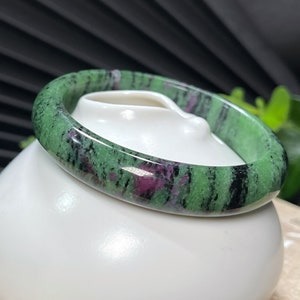 Puede incluir: Un brazalete de piedra preciosa verde, negro y rosa. El brazalete est&aacute; hecho de una piedra lisa y pulida y tiene un aspecto natural y org&aacute;nico.