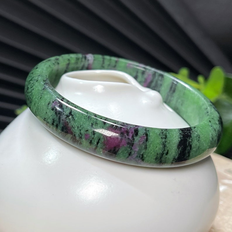 Puede incluir: Un brazalete de piedra preciosa verde, negro y rosa. El brazalete est&aacute; hecho de una piedra lisa y pulida y tiene un aspecto natural y org&aacute;nico.