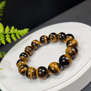 Pode incluir: Uma pulseira com contas redondas e polidas de olho de tigre. As contas são marrom dourado e marrom escuro, com um acabamento brilhante. A pulseira é exibida em uma superfície branca.
