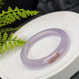 Może przedstawiać: Gładka, półprzezroczysta bransoletka typu bangle w kolorze lawendowym z polerowanym wykończeniem. Okrągła obręcz posiada małą, wyraźną inkluzję czerwonobrązowych, nakrapianych osadów mineralnych zatopionych w jasnofioletowym kamieniu.