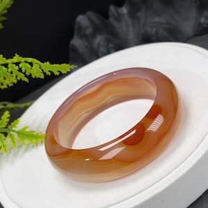 Può includere: Un bracciale rigido liscio e levigato, realizzato in pietra agata traslucida. Il bracciale presenta una calda tavolozza di colori a bande, con sfumature di arancione intenso, terra di Siena bruciata e bianco crema, con motivi naturali.
