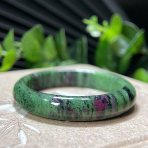 Könnte beinhalten: Ein grünes und rosafarbenes Edelstein-Armband. Das Armband ist aus poliertem Stein gefertigt und hat eine glatte, abgerundete Oberfläche.