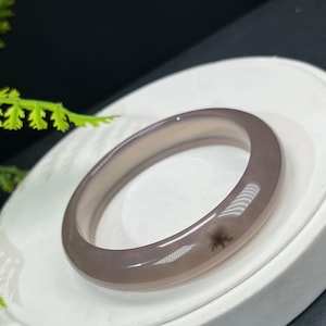 Può includere: Un braccialetto a cerchio in pietra preziosa liscia, grigio-viola. Il braccialetto ha un design circolare e una finitura lucida. Il colore è una tonalità tenue e smorzata di grigio-viola con sottili variazioni. Il braccialetto è esposto su una superficie bianca.