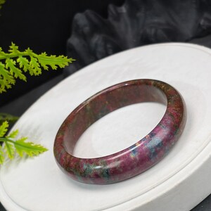 May include: A polished bangle bracelet crafted from a smooth, rounded stone featuring a mottled pattern of deep magenta, dark red, and teal blue hues. The surface of the circular jewellery piece has a glossy finish that reflects light across its variegated colours.