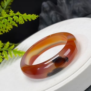 May include: A smooth, polished bangle bracelet crafted from translucent orange and red agate stone. The circular band features natural color variations, including deep amber bands and dark, moss-like inclusions visible within the polished surface.
