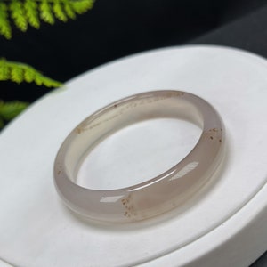 May include: A light gray agate bangle bracelet with brown flecks. The bangle is smooth and polished.