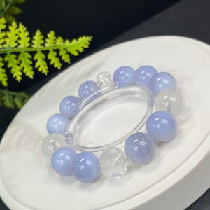 Puede incluir: Un brazalete con cuentas esféricas de color azul claro y transparentes. Las cuentas están ensartadas en una banda circular transparente. El brazalete se exhibe sobre una superficie blanca y circular, con un fondo oscuro.