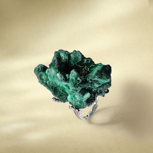 May include: A ring featuring a large, green malachite stone set in a silver band. The stone has a rough, natural texture and a unique, organic shape.