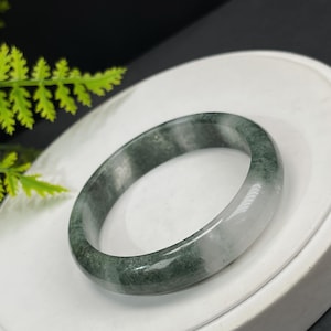 Może przedstawiać: Jadeitowa bransoletka z gładką, wypolerowaną powierzchnią. Bransoletka charakteryzuje się połączeniem zielonych i białych odcieni, tworząc efekt marmuru. Jadeit umieszczono na białym tle, podkreślając jego naturalne kolory i faktury.