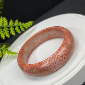 Op de afbeelding: Een koraalkleurige armband met een gespikkeld patroon. De armband is een massieve band van gepolijste steen, met kleurvariaties van licht tot donker koraal. De armband wordt gepresenteerd op een wit oppervlak.