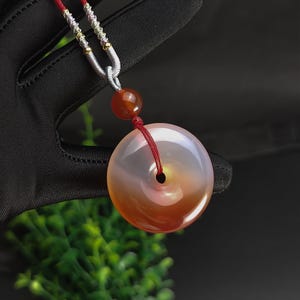 May include: A pendant necklace featuring a large, round, polished stone with a central hole. The stone has a gradient of colors, from clear to orange-red. The necklace has a red cord and a silver-colored chain.