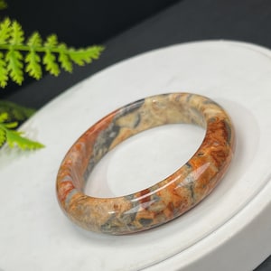 May include: A polished, oval-shaped bangle bracelet. The bracelet features a blend of colors, including shades of orange, brown, gray, and beige. The stone-like material has a marbled appearance. The bracelet is a jewelry item.
