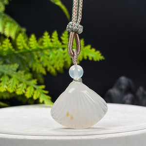 May include: A translucent white shell-shaped pendant carved from stone, featuring subtle vertical ridges and two small yellow spots. The pendant is suspended from a brown and green braided cord, accented by a single smooth, round white bead.