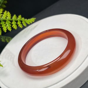 May include: A polished, translucent orange-red bangle bracelet. The smooth, circular bracelet is made of a gemstone material and rests on a white surface. The bangle is a jewelry item.