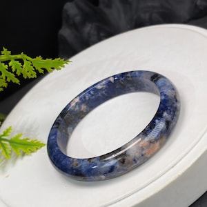 May include: A polished bangle bracelet crafted from natural stone, featuring a smooth, rounded surface with a mottled pattern of deep blue, black, and soft peach-colored inclusions throughout the material, showcasing a glossy, reflective finish.