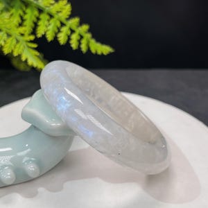 以下が含まれることがあります： 虹色の光沢を持つ白い翡翠のバングル。バングルは滑らかで磨かれており、わずかに丸みを帯びた形をしています。