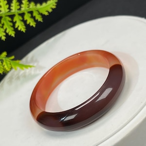 May include: A polished, oval-shaped bangle bracelet. The bracelet transitions from a light orange hue to a deep burgundy color. The smooth surface reflects light, highlighting the natural variations in the stone. A piece of jewelry.