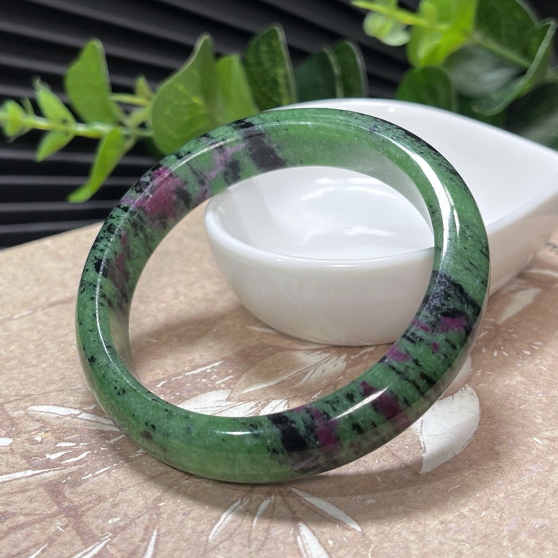 Puede incluir: Un brazalete de jade verde, negro y rosa. El brazalete es liso y pulido.