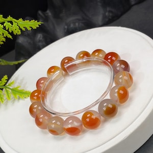 Könnte beinhalten: Ein Armband mit orangefarbenen und weißen Achatperlen. Die runden, polierten Perlen haben ein durchscheinendes, gebändertes Aussehen. Das Armband wird von einem klaren, gebogenen Kunststoffteil zusammengehalten. Die Perlen haben einen Durchmesser von etwa 1 cm.