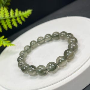Peut inclure: Un bracelet composé de perles rondes gris-vert translucides. Les perles sont enfilées ensemble, formant un cercle. Le bracelet est présenté sur une surface blanche, avec un fond sombre. Chaque perle mesure environ 1 cm de diamètre.