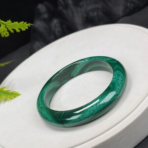 May include: A polished, oval-shaped malachite bracelet. The bracelet features a vibrant green color with darker green and brown swirling patterns. The gemstone bracelet is displayed on a white surface.