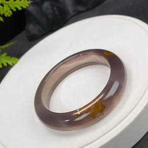 以下が含まれることがあります： 紫色の半透明のバングルブレスレット。 磨かれた表面には、薄茶色と黄色のインクルージョンがあります。 ブレスレットは円形で、石または同様の素材で作られているようです。