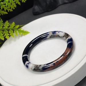 May include: A polished bangle bracelet crafted from a smooth, rounded stone featuring a marbled pattern of deep navy blue, white, and earthy brown tones. The circular accessory has a glossy, reflective surface that highlights the natural mineral variations.