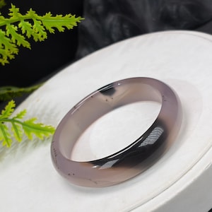 May include: A smooth, translucent grey agate bangle bracelet with subtle dark inclusions and a polished finish. The circular stone band features a rounded profile and a consistent thickness, showcasing natural variations in the grey mineral structure.