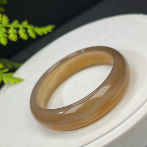 May include: A polished, light brown gemstone bangle bracelet. The bracelet has a smooth, rounded design and a translucent appearance. The color transitions from a light tan to a darker brown. The bracelet is displayed on a white surface.