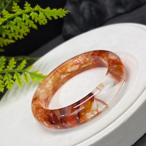 May include: A smooth, circular bangle bracelet crafted from clear, polished material featuring internal inclusions of deep red, orange, and golden-brown hues that create a swirling, marbled effect throughout the translucent structure.