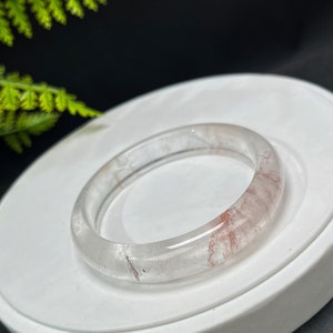Op de afbeelding: Een heldere kwarts armband met rode en witte insluitsels. De armband is glad en gepolijst.